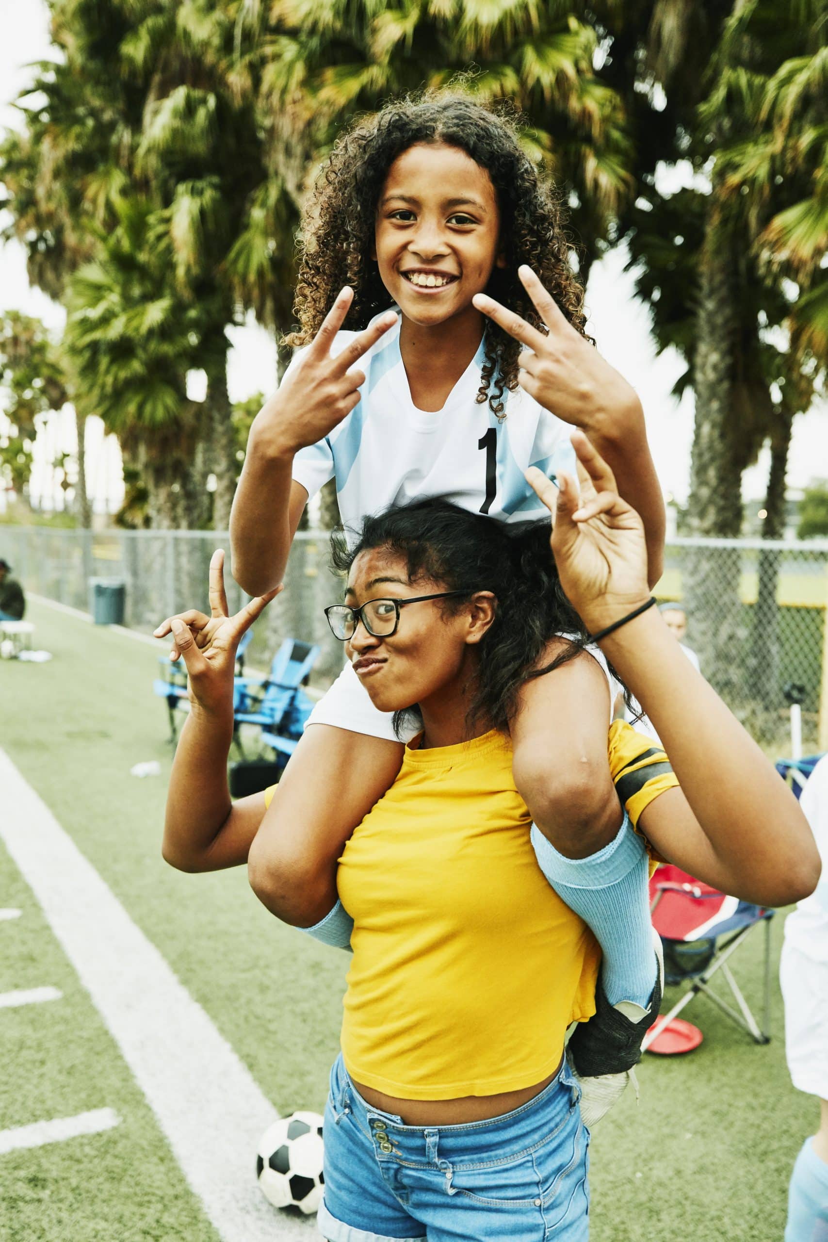 One girl sitting on another girl's shoulders laughing and making peace signs