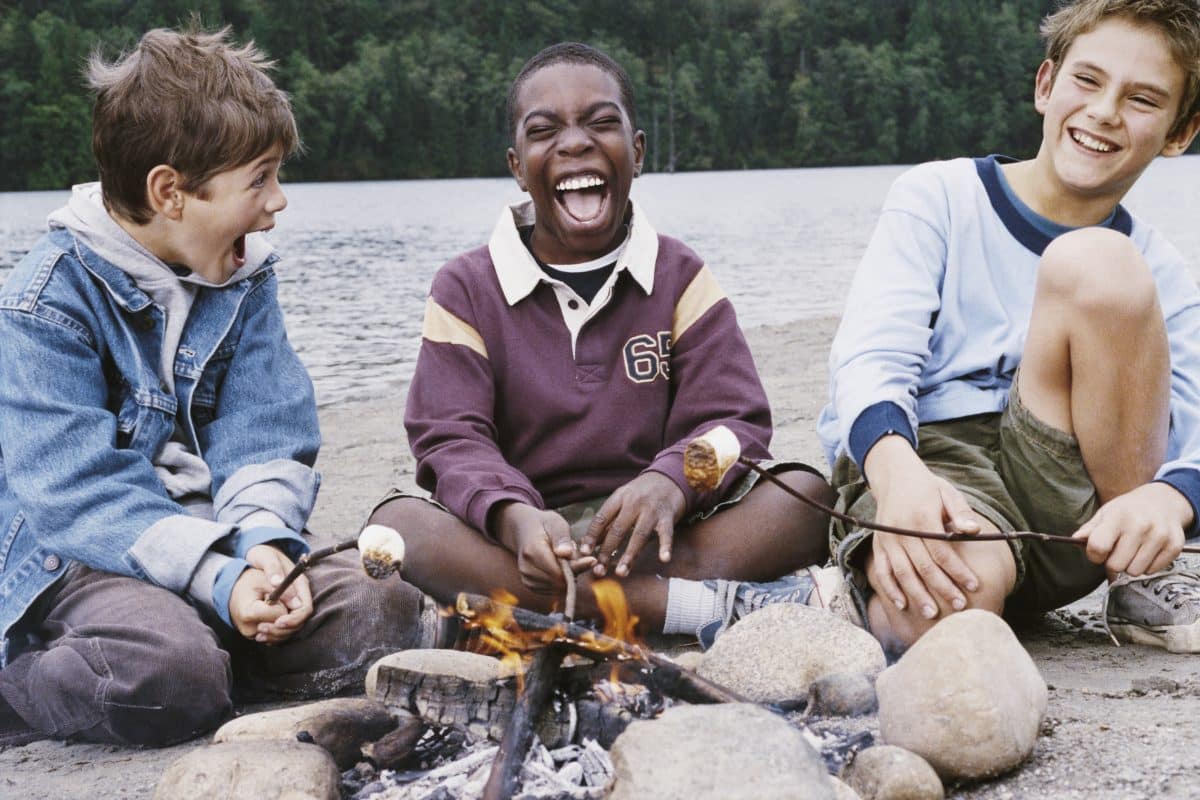 Three boys sitting around a campfire making s'mores text to a lake and laughing